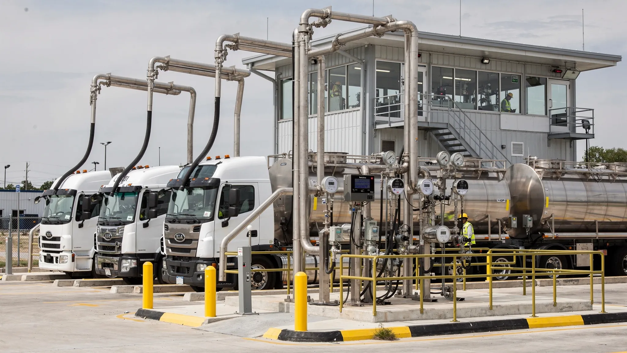 A fuel terminal rack with multiple tanker trucks loading gasoline and diesel, visible meters and hoses, and a control building in the background. The scene emphasizes high-volume fuel distribution and measurement at the rack.